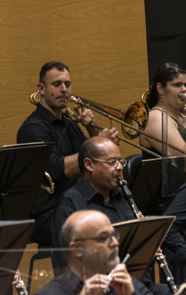 Vemos parte dos músicos da orquestra tocando trompetes e outros instrumentos de sopro.