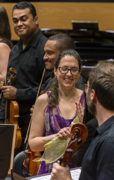 Vemos a solista Brigitta Calloni sorrindo, segurando um buquê de flores. Ao seu redor é possível ver parte da orquestra.