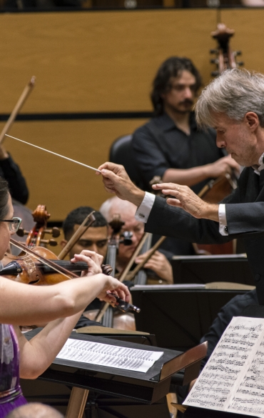 Vemos o maestro Pawet Przytocki e a solista Brigitta Calloni. Ela está à esquerda da imagem, tocando violino, o maestro está à direita, um pouco mais alto que a solista, com a batuta em mãos.