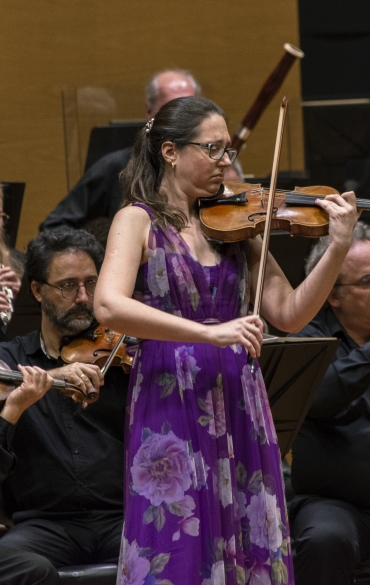 Vemos no centro a solista Brigitta Calloni tocando violino e uma parte da orquestra atrás dela.