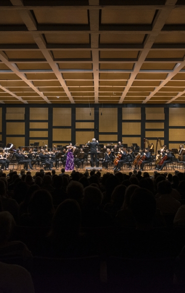 Vemos todo o palco da Sala Sinfônica da OSPA. É possível ver toda a orquestra, à sua frente o maestro e a solista, e uma parte do público de costas.