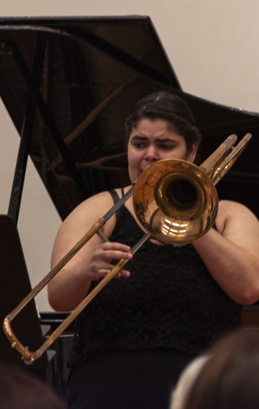 Vemos a trombonista Sabryna Pinheiro tocando, atrás dela vemos parte do piano.