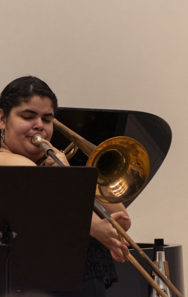 Vemos a trombonista Sabryna Pinheiro tocando, atrás dela vemos o piano e à sua frente vemos parte do suporte de partitura.