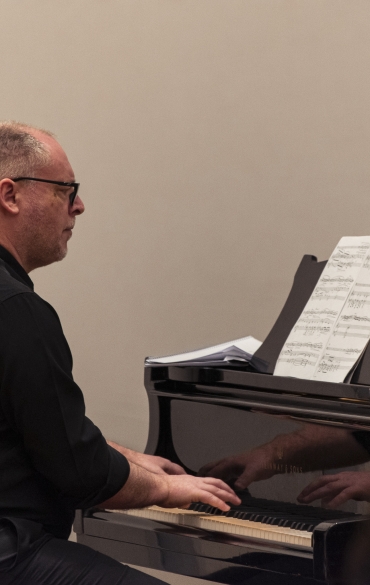 Vemos o pianista Paulo Bergmann tocando. Ele lê a partitura à sua frente, apoiada em cima do piano.