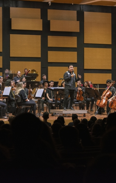 Vemos todo o palco da Sala Sinfônica. Toda a OSPA Jovem distribuída no palco, formada principalmente por adolescentes, à sua frente o maestro Arthur Barbosa, no centro, e à direita da imagem o mestre de cerimônias Oséas Duarte.