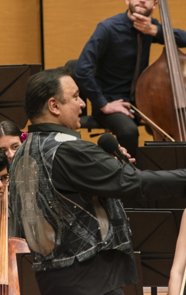 Vemos no centro o maestro Arthur Barbosa segurando a sua batuta. Ele está de lado, levemente inclinado para trás, ao fundo vemos alguns dos jovens da orquestra.