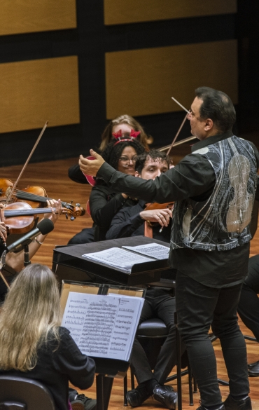 Vemos, à direita, o maestro Arthur Barbosa com os braços abertos e sua batuta, regendo a orquestra. À esquerda é possível ver parte dos jovens da orquestra, a maioria com violinos.