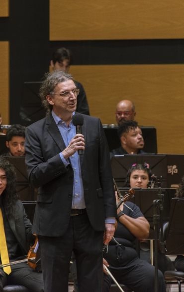 No centro da imagem, vemos o diretor da Escola de Música da OSPA, Diego Grendene. Ele segura um microfone e fala com a plateia. Atrás dele vemos alguns dos jovens da OSPA Jovem, alguns deles usam fantasias como gravatas e tiaras.