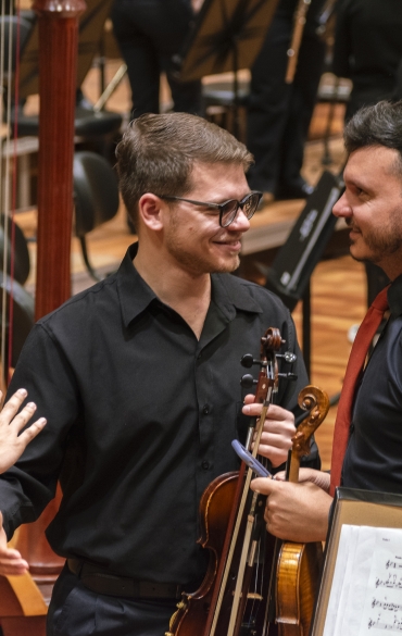 No centro da imagem vemos um violinista sorrindo. Ele olha para a direita e levanta levemente a mão para um cumprimentar o músico à sua frente.