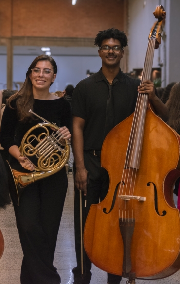 Vemos quatro jovens da OSPA Jovem posando para a foto. Da esquerda para a direita temos uma menina segurando um violoncelo, uma menina segurando uma trompa, um menino segurando um contrabaixo e uma menina segurando um fagote. Todos sorriem.