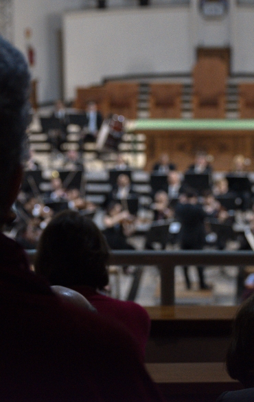 Vemos algumas pessoas do público, de costas, olhando para a orquestra.