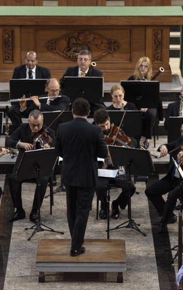 Vemos boa parte da orquestra e à sua frente o maestro Evandro Matté, de costas para a câmera.