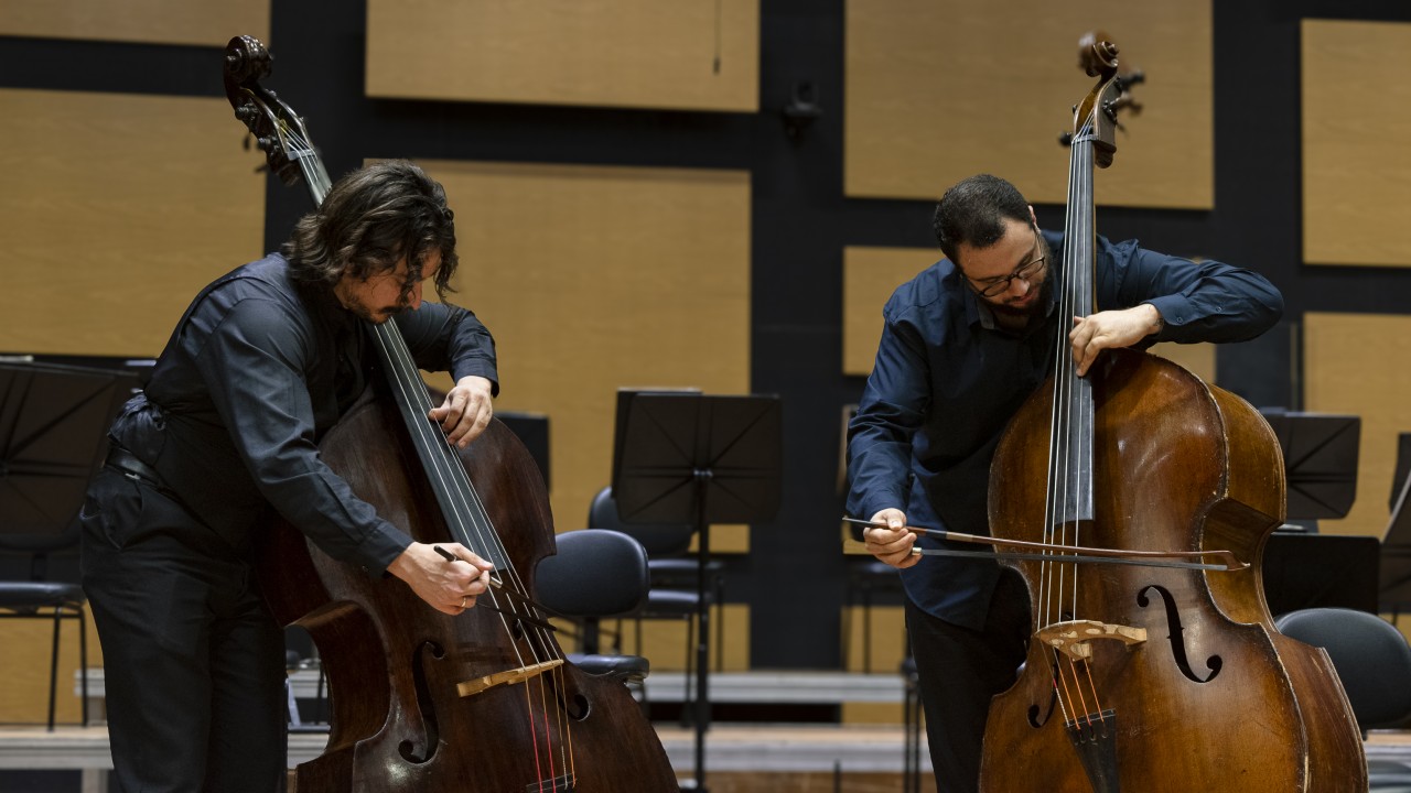 Foto horizontal de dois homens tocando contrabaixos em uma sala de concertos. Ambos usam camisas escuras e seguram arcos. O homem à esquerda inclina-se levemente para a frente enquanto toca o instrumento, que está à sua esquerda. O homem à direita também inclina o corpo e o arco para a frente ao tocar. Ao fundo, há cadeiras e estantes de partituras em frente a painéis de madeira retangulares em tons neutros.