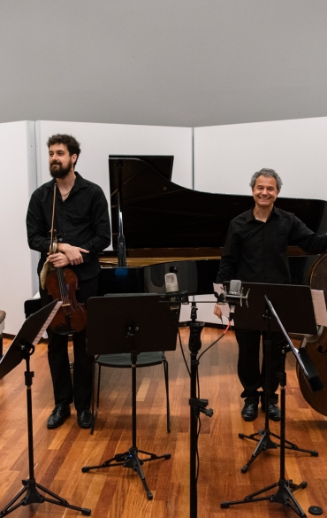 Grupo de cinco pessoas em pé com instrumentos. O fundo é branco e as pessoas estão vestidas com roupas escuras. Entre os instrumentos, estão: piano, contrabaixo, violino, viola e violoncelo.