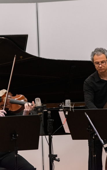 Fundo branco, com piano de fundo e dois homens a frente, um executando uma música na viola ao lado de outro homem que está tocando no contrabaixo.