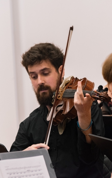 Fundo branco com homem centralizado tocando viola. Ao fundo, há o rosto de uma mulher de óculos.