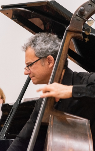 A imagem tem fundo branco, no plano mais ao fundo há uma mulher tocando piano e, a frente, um homem tocando contrabaixo.