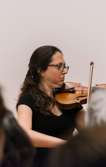 Mulher tocando violino no centro, há um fundo branco e aparece parte da plateia.