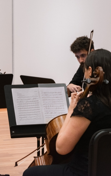 Grupo de cinco pessoas em uma sala, no plano a frente há uma mulher de costas, ao seu lado há dois homens com instrumentos, ao fundo, do lado direito, há uma mulher no piano e, ao lado esquerdo, outra mulher com violino.