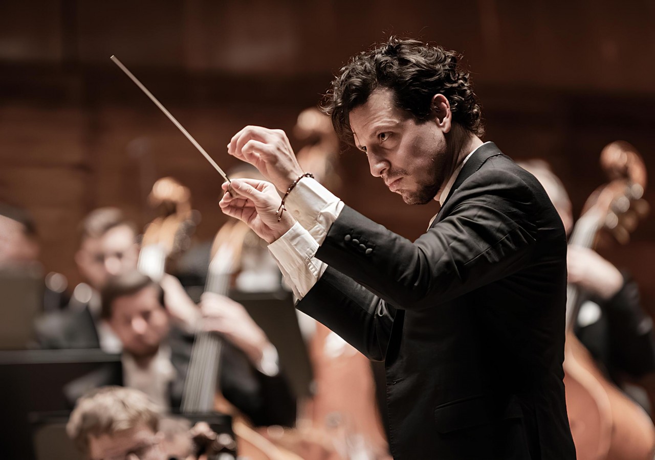 A imagem mostra um maestro em ação durante uma performance orquestral. Ele está em destaque, de perfil, segurando uma batuta com ambas as mãos, com expressão séria e concentrada. O maestro veste um terno preto com camisa branca, e seus movimentos parecem elegantes e precisos, dirigindo a orquestra com intensidade. Ao fundo, desfocados, aparecem músicos e seus instrumentos, como violoncelos e contrabaixos, reforçando o ambiente orquestral. A iluminação quente do local realça os tons de marrom e bege, criando uma atmosfera sofisticada e intimista.