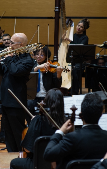 
Esta imagem mostra um trombonista em destaque no centro, tocando de frente para a orquestra e com o trombone erguido. O músico está de pé em uma plataforma à frente de outros instrumentistas. Atrás dele, os demais músicos, incluindo violinistas, pianistas e uma harpista, observam com atenção, alguns com expressões de aprovação e outros com sorrisos. A harpista, à direita, olha com expressão de apreciação e calma para a performance.

A orquestra está em um palco de madeira, e todos os músicos estão vestidos de maneira formal, em trajes escuros. As partituras e instrumentos em mãos sugerem uma apresentação ou ensaio formal. A disposição dos músicos em semicírculo e a interação entre eles refletem a sinergia e a harmonia típicas de uma orquestra bem coordenada. O ambiente geral é de concentração e apreciação musical.