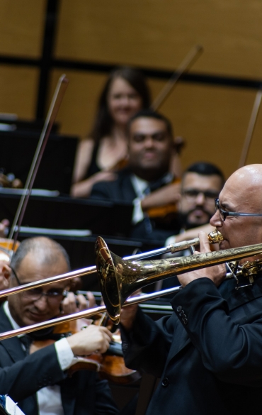 Nesta imagem, o trombonista aparece novamente em destaque, tocando seu instrumento com grande concentração e intensidade. Ao fundo, músicos da orquestra, especialmente violinistas, acompanham a performance com expressões de alegria e apreciação. Alguns deles sorriem, o que sugere uma conexão positiva e uma atmosfera de entusiasmo durante a apresentação.

A cena captura a interação entre o trombonista e os outros músicos, enfatizando o espírito colaborativo da orquestra. Todos os músicos estão vestidos formalmente em trajes escuros, e a expressão dos ouvintes reflete um momento de harmonia e entusiasmo musical. A imagem transmite uma energia vibrante e a sinergia entre os instrumentistas no palco.