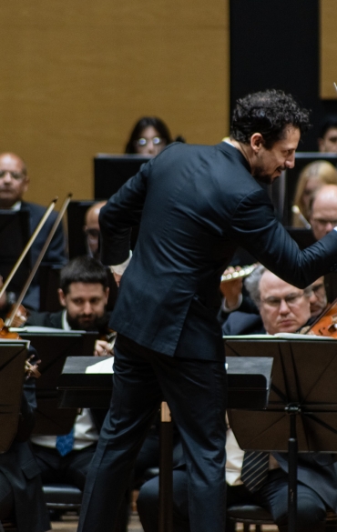 A imagem mostra um maestro no centro, conduzindo uma orquestra. Ele está de costas para a câmera e usa um traje formal preto. Sua mão direita está levantada, segurando uma batuta, e ele parece estar concentrado na condução dos músicos à sua frente. Ao redor dele, músicos de orquestra tocam instrumentos de corda, como violinos e violas, todos vestidos formalmente. O fundo é neutro, destacando o maestro e os músicos na execução. A expressão dos músicos é focada, refletindo a concentração durante a performance.