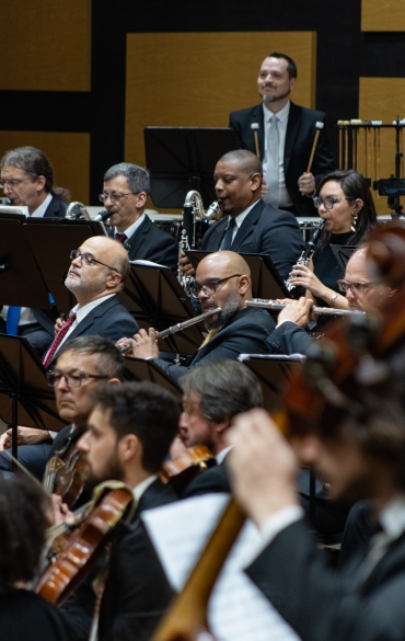 
A imagem apresenta um grupo de músicos de uma orquestra durante uma apresentação. Eles estão em fileiras, com foco em instrumentos de sopro, como flautas, clarinetes e trompas, além de alguns músicos nas seções de cordas, como violinos e violas, na frente. Todos estão vestidos formalmente e parecem concentrados em suas partituras ou instrumentos. Ao fundo, vemos percussionistas e outros músicos. A atmosfera é de concentração e profissionalismo, refletindo o ambiente de uma apresentação orquestral.