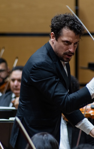 A imagem mostra um maestro conduzindo uma orquestra, com uma expressão intensa e concentrada, evidenciando a sua conexão com a música. Ele segura a batuta com uma mão levantada, indicando o tempo e a dinâmica para os músicos à sua frente. No fundo, é possível ver os violinistas focados em suas partituras, acompanhando a regência do maestro. A cena transmite o poder e a emoção de uma performance musical ao vivo, destacando o papel do maestro em guiar a interpretação coletiva da orquestra.