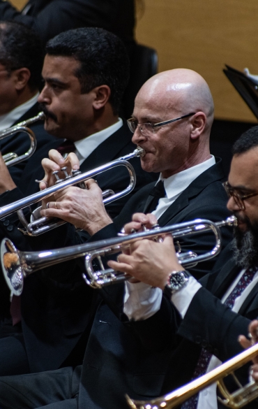 
A imagem mostra uma seção de trompetistas em uma orquestra. Cinco músicos tocam seus instrumentos, posicionados em uma fileira. Todos usam ternos escuros, camisas brancas e gravatas escuras. Eles estão concentrados, com os lábios pressionados contra os bocais dos trompetes. Um dos músicos é careca e usa óculos, enquanto outros têm cabelos e barbas de diferentes estilos. Atrás deles, é possível ver partes de outros instrumentos, como trombones e partituras, sugerindo que eles estão em uma performance ao vivo em um palco.