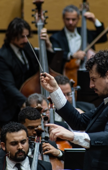 
A imagem mostra uma orquestra em performance, com foco nos músicos da seção de cordas, especialmente nos contrabaixistas e violoncelistas. No primeiro plano, um maestro com expressão concentrada conduz a orquestra, segurando uma batuta na mão direita e mantendo a outra mão levantada. Ele está vestindo um terno escuro. Atrás dele, vários músicos tocam seus instrumentos, todos também usando roupas formais. Ao fundo, quatro contrabaixistas estão posicionados, com seus arcos nas cordas e expressões focadas. A atmosfera é de intensa concentração, típica de uma apresentação ao vivo em um auditório.