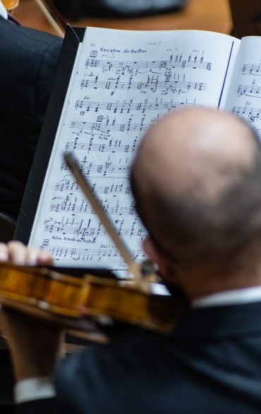 A foto mostra um músico de orquestra visto de costas, segurando um violino e olhando para uma partitura aberta à sua frente. A partitura exibe várias notas musicais organizadas em linhas, indicando uma composição complexa. O músico, que tem cabeça calva, está vestido formalmente com um terno preto. A imagem parece capturar um momento de concentração enquanto ele se prepara para tocar, com o violino e o arco prontos em suas mãos. O ambiente é iluminado e formal, sugerindo uma performance ou ensaio profissional em um auditório.