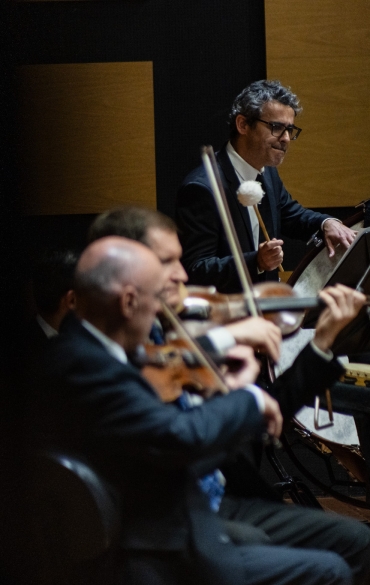 A imagem mostra uma apresentação de uma orquestra sinfônica. No primeiro plano, há músicos tocando instrumentos de cordas, provavelmente violinos e violas, todos concentrados em suas partituras. Eles estão vestidos formalmente, com trajes escuros. No plano mais ao fundo, um percussionista está de pé, segurando uma baqueta com uma grande cabeça acolchoada, prestes a tocar um bumbo. Ele também está vestindo terno e usa óculos. A iluminação é suave e o ambiente parece elegante, com painéis de madeira ao fundo. O foco da imagem está no músico de percussão, mas a composição também revela a intensidade e a concentração dos outros músicos ao redor.