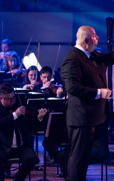 A imagem mostra um maestro regendo uma orquestra durante uma apresentação. Ele está de pé em frente à orquestra, com uma das mãos levantada, gesticulando para os músicos. Atrás dele, vários instrumentistas, principalmente violinistas, tocam atentamente, seguindo suas instruções. A iluminação é azulada, criando uma atmosfera dramática e intensa. O ambiente sugere uma apresentação formal, com os músicos concentrados em seus instrumentos e partituras, enquanto o maestro conduz a performance com precisão e autoridade.