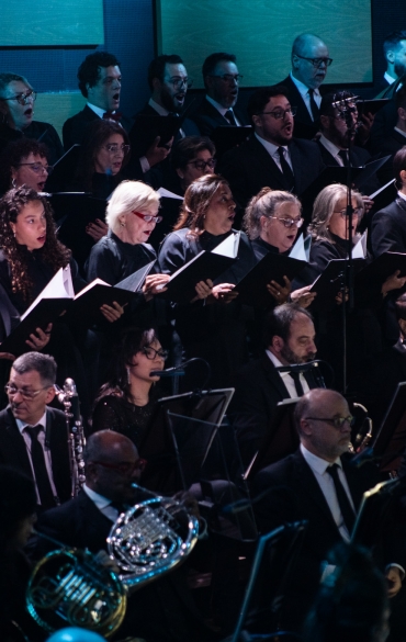 
A imagem mostra um coral e uma orquestra sinfônica durante uma apresentação. Os cantores do coro estão dispostos em fileiras no fundo, todos vestidos com roupas formais escuras e segurando partituras. Eles aparentam estar concentrados, com a maioria deles cantando de forma expressiva.

Na frente do coral, vemos parte da orquestra, composta por músicos tocando instrumentos de sopro, como trompas e clarinetes. Eles também estão vestidos formalmente. O ambiente tem uma iluminação suave e escura, com alguns focos de luz sobre os integrantes, destacando seus rostos e instrumentos. A atmosfera parece ser solene e dedicada à música clássica, criando um clima de envolvimento e intensidade musical.