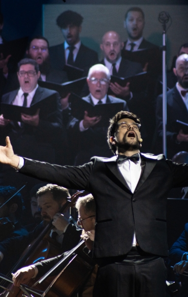 
A imagem mostra um coral e uma orquestra sinfônica durante uma apresentação. Os cantores do coro estão dispostos em fileiras no fundo, todos vestidos com roupas formais escuras e segurando partituras. Eles aparentam estar concentrados, com a maioria deles cantando de forma expressiva.

Na frente do coral, vemos parte da orquestra, composta por músicos tocando instrumentos de sopro, como trompas e clarinetes. Eles também estão vestidos formalmente. O ambiente tem uma iluminação suave e escura, com alguns focos de luz sobre os integrantes, destacando seus rostos e instrumentos. A atmosfera parece ser solene e dedicada à música clássica, criando um clima de envolvimento e intensidade musical.