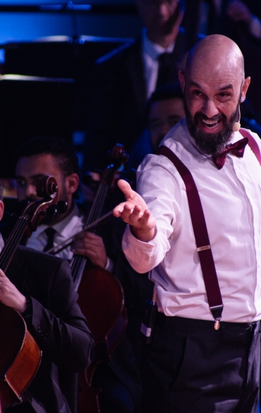 A imagem mostra um homem de meia-idade, careca e com barba, realizando uma performance teatral durante uma apresentação musical. Ele veste uma camisa branca, suspensórios vermelhos e uma gravata borboleta, e segura um candelabro dourado com velas brancas, que parece fazer parte do figurino ou do cenário. O homem está voltado para outra pessoa de costas, que usa um vestido de alças, provavelmente uma personagem da cena.

Sua expressão facial é expressiva, como se estivesse encenando ou falando algo com intensidade. Ao fundo, músicos da orquestra, incluindo um violoncelista, observam a cena, e o ambiente está iluminado com tons de azul, criando uma atmosfera dramática e envolvente.