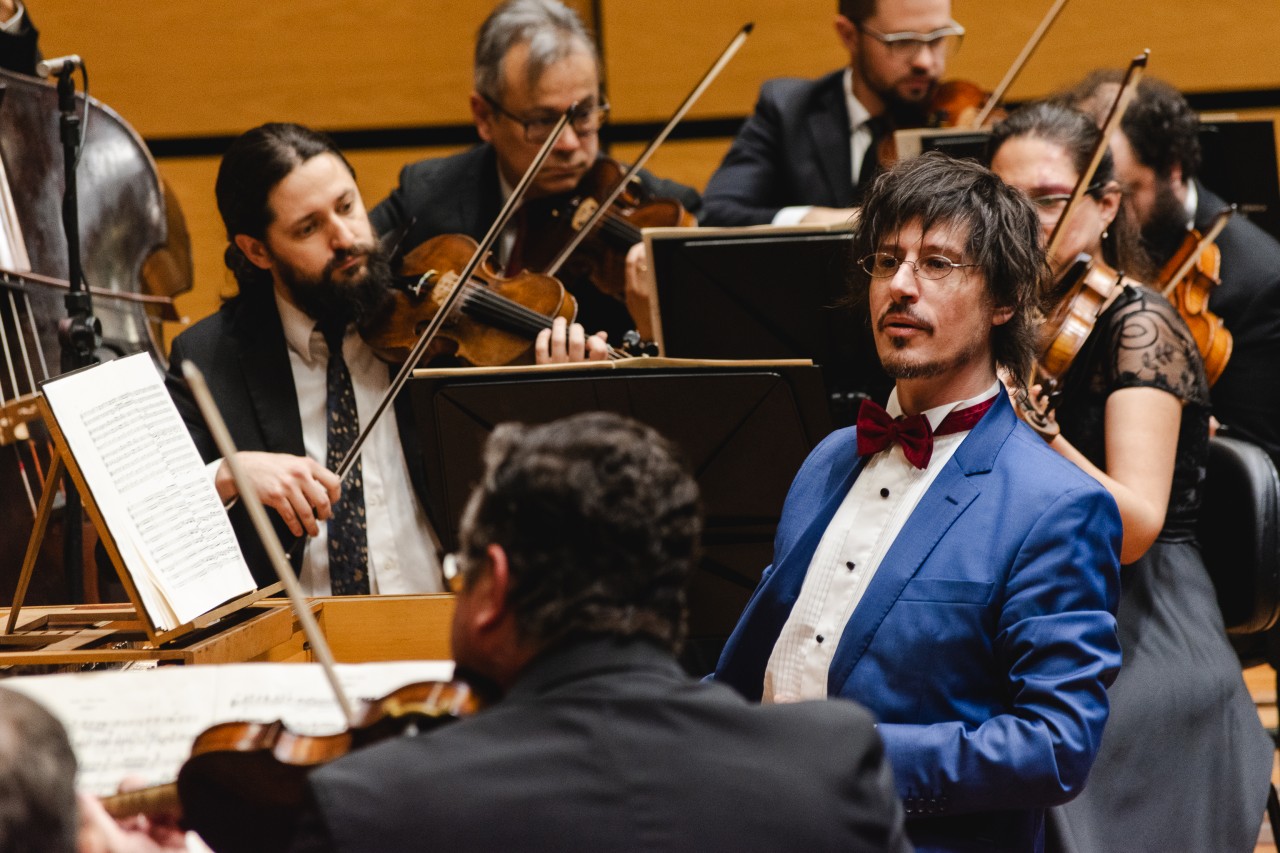 A imagem mostra uma orquestra durante uma apresentação. No centro, está o maestro, vestindo um terno azul vibrante, camisa branca e uma gravata borboleta vermelha, com uma expressão focada, direcionando o olhar para a orquestra. Ele usa óculos e tem cabelo escuro e bagunçado.

Ao seu redor, diversos músicos tocam violinos e outros instrumentos de corda, todos usando roupas de concerto formais, como ternos escuros e vestidos pretos. O ambiente é uma sala de concertos com painéis de madeira clara ao fundo. Partituras abertas estão dispostas em suportes à frente dos músicos, que seguem concentrados na execução da peça.