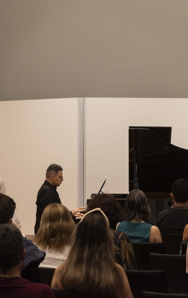 A imagem mostra uma apresentação musical em um espaço fechado. Um homem, de perfil, está sentado ao piano, tocando a peça, enquanto o público, composto por várias pessoas sentadas em cadeiras, observa. O piano de cauda preto está posicionado à direita da imagem, com uma estante de partitura ao lado. Ao fundo, há divisórias brancas simples, que formam o cenário. O ambiente parece ser uma sala de concertos ou espaço cultural, com iluminação neutra.