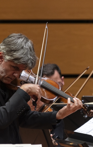 A imagem mostra um violinista tocando durante uma performance orquestral. Ele está vestido formalmente com um terno preto, concentrado enquanto toca o violino. No fundo, outros músicos e instrumentos da orquestra são parcialmente visíveis, assim como partituras. A parede ao fundo é lisa, com tom bege, típico de um auditório ou sala de concerto.