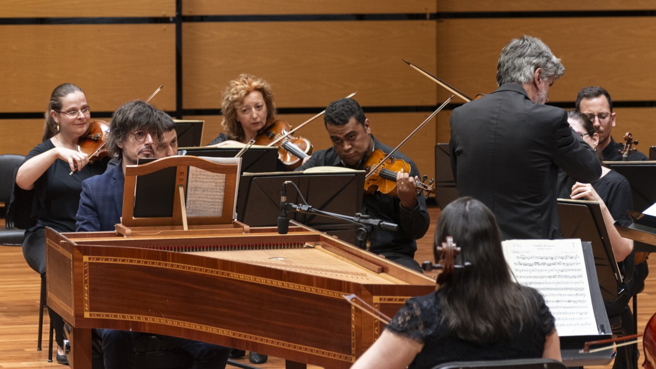 
A imagem apresenta uma performance musical em um palco, com músicos tocando instrumentos clássicos. No centro está um cravo em madeira detalhada, um instrumento típico de música barroca, com partituras abertas sobre ele. Atrás do cravo, violinistas e outros instrumentistas da orquestra aparecem concentrados, com expressões sérias. O ambiente é um auditório de madeira clara, que proporciona uma atmosfera acolhedora e acústica ideal para esse tipo de música. A disposição dos músicos e a escolha dos instrumentos sugerem a execução de uma obra do período barroco.