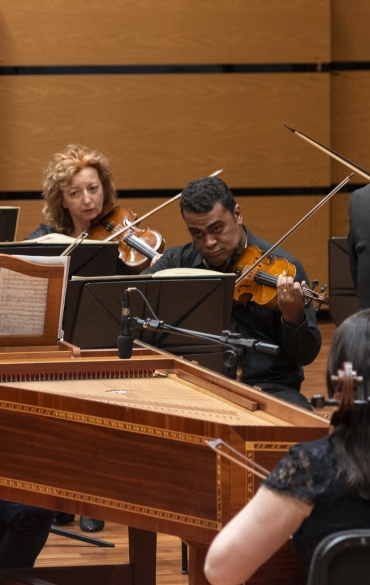 
A imagem apresenta uma performance musical em um palco, com músicos tocando instrumentos clássicos. No centro está um cravo em madeira detalhada, um instrumento típico de música barroca, com partituras abertas sobre ele. Atrás do cravo, violinistas e outros instrumentistas da orquestra aparecem concentrados, com expressões sérias. O ambiente é um auditório de madeira clara, que proporciona uma atmosfera acolhedora e acústica ideal para esse tipo de música. A disposição dos músicos e a escolha dos instrumentos sugerem a execução de uma obra do período barroco.