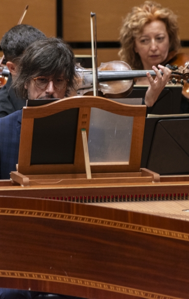 
A imagem mostra um músico tocando cravo em destaque, com um grupo de violinistas ao fundo. O cravista, com cabelos compridos e óculos, está concentrado em sua performance. O cravo, feito em madeira ornamentada, ocupa uma posição central e é um instrumento típico da música barroca. Atrás dele, os violinistas estão posicionados, segurando seus instrumentos e acompanhando a música. O ambiente é formal, provavelmente um auditório de concerto com painéis de madeira clara, e o clima geral transmite uma atmosfera de dedicação e apreço à música clássica.