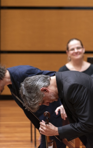 A imagem mostra dois músicos em um palco de madeira, inclinando-se para frente em uma reverência, possivelmente ao final de uma apresentação. Ambos usam ternos escuros. Um deles segura um instrumento de cordas, aparentemente um violino ou viola. Ao fundo, outros músicos estão sentados, sorrindo, com um cravo parcialmente visível. A parede ao fundo tem um padrão liso e tons de madeira clara.