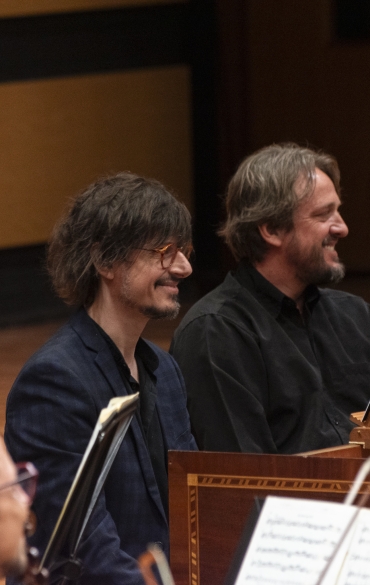 A imagem mostra músicos sentados em um palco de madeira. À esquerda, um violinista segura seu instrumento. No centro, dois homens sorriem: o primeiro veste um terno azul escuro e usa óculos de armação laranja; o segundo, com cabelos grisalhos, veste uma camisa preta. À direita, outros músicos aparecem parcialmente, com partituras e um cravo visíveis. O fundo tem uma parede de madeira clara com detalhes escuros. A atmosfera sugere uma apresentação de música clássica.