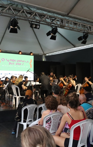 A imagem mostra uma apresentação musical em um palco coberto por uma estrutura de lona branca. Um grupo de músicos, incluindo violinistas e violoncelistas, está tocando sob a orientação de um maestro ao centro. Os músicos estão sentados em cadeiras brancas, com partituras em suportes à frente. À direita do palco, há duas faixas com a mensagem "O Brasil é a nossa energia" e a marca da Petrobras, além de ilustrações relacionadas ao tema.

No fundo do palco, há um telão exibindo o texto "O tempo passa por aqui", com detalhes em cores vibrantes. O público, composto por pessoas de diferentes idades, está sentado em cadeiras de plástico brancas, assistindo atentamente à apresentação. A iluminação do palco é feita por refletores fixados em uma estrutura metálica suspensa. O ambiente sugere um evento cultural com apoio de patrocinadores.