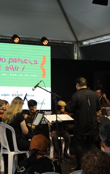 A imagem mostra uma apresentação musical em um palco coberto, com uma orquestra de músicos jovens tocando instrumentos de cordas, como violinos, violoncelos e contrabaixos. À frente, um maestro conduz o grupo, vestindo roupas pretas e posicionado de costas para o público. O fundo apresenta um telão com fundo verde e a mensagem: "O tempo passe, a por aqui", seguida da identificação do evento como "70ª Feira do Livro de Porto Alegre" e datas: "De 01 a 20/11 das 10 às 20h". A parte inferior do telão exibe logos de apoiadores e patrocinadores. A plateia está parcialmente visível na parte inferior da imagem, sentada em cadeiras brancas de plástico.