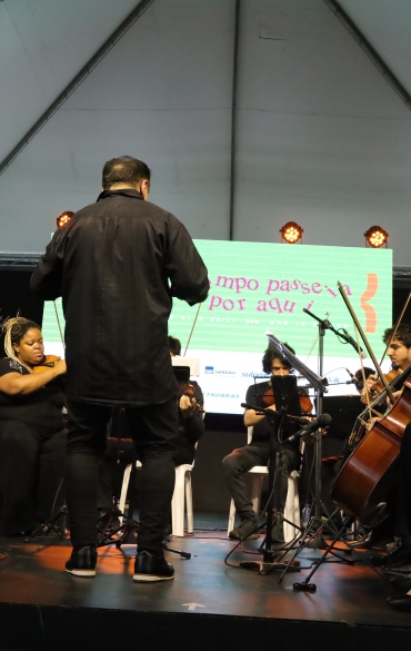 
A imagem retrata uma orquestra se apresentando em um palco sob uma estrutura coberta. Os músicos, sentados em cadeiras brancas, estão vestidos predominantemente de preto e tocam instrumentos de cordas, como violinos, violoncelos e contrabaixos. À frente do grupo, o maestro conduz a performance, de costas para a câmera, vestindo uma roupa preta. À esquerda, há um microfone posicionado em um pedestal próximo aos músicos.

Ao fundo, um grande telão verde exibe o texto: "O tempo passe, a por aqui", junto com a identificação do evento "70ª Feira do Livro de Porto Alegre" e informações sobre datas e horários: "De 01 a 20/11 das 10 às 20h". Abaixo do texto, diversos logotipos de patrocinadores e apoiadores são visíveis. A iluminação do palco inclui refletores, criando uma atmosfera acolhedora para o público.