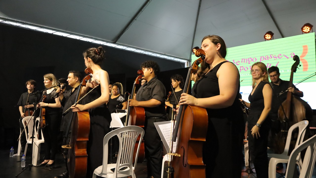 A imagem mostra os músicos de uma orquestra em pé no palco, segurando seus instrumentos, como violinos, violoncelos e contrabaixos, em um momento de agradecimento ao público. Eles vestem roupas pretas formais e estão posicionados em frente a cadeiras brancas. Ao centro, o maestro também participa do momento, segurando um microfone, aparentemente falando ou agradecendo.

O fundo exibe novamente o grande telão com fundo verde, onde está escrito: "O tempo passe, a por aqui", seguido da identificação do evento "70ª Feira do Livro de Porto Alegre". A iluminação do palco destaca os músicos, enquanto o teto coberto sugere um evento em espaço semiaberto. Os semblantes dos músicos expressam seriedade e concentração, enquanto o público não aparece diretamente na cena.