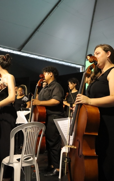 A imagem mostra os músicos de uma orquestra em pé no palco, segurando seus instrumentos, como violinos, violoncelos e contrabaixos, em um momento de agradecimento ao público. Eles vestem roupas pretas formais e estão posicionados em frente a cadeiras brancas. Ao centro, o maestro também participa do momento, segurando um microfone, aparentemente falando ou agradecendo.

O fundo exibe novamente o grande telão com fundo verde, onde está escrito: "O tempo passe, a por aqui", seguido da identificação do evento "70ª Feira do Livro de Porto Alegre". A iluminação do palco destaca os músicos, enquanto o teto coberto sugere um evento em espaço semiaberto. Os semblantes dos músicos expressam seriedade e concentração, enquanto o público não aparece diretamente na cena.